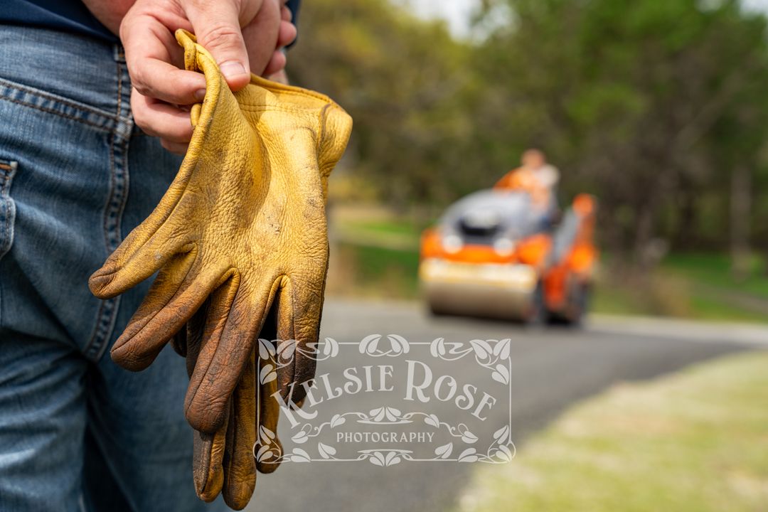 Close-up of yellow worn leather work gloves held by a worker with orange road roller in background.