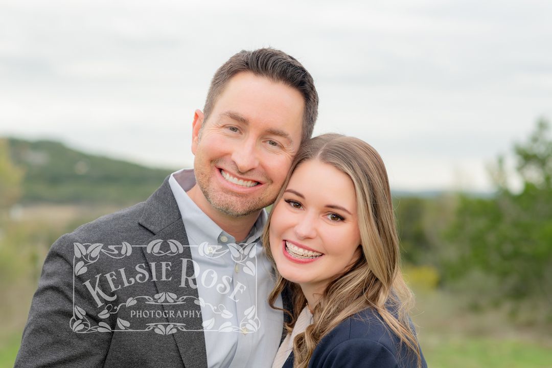 Smiling couple posing cheek to cheek outdoors with green hills in background, professional portrait photography.