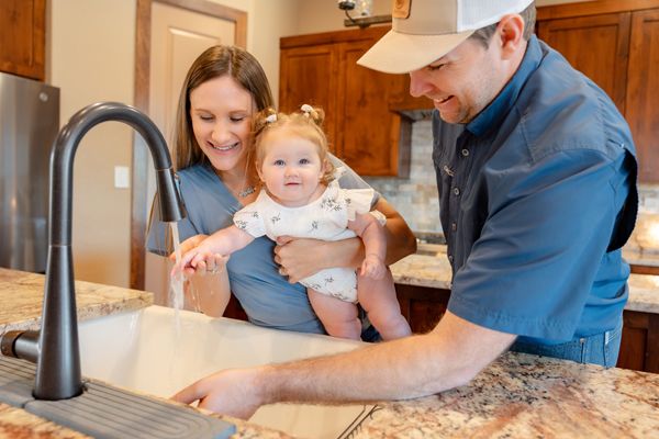 Couple with smiling baby interact with water flowing from their kitchen faucet in modern kitchen with granite countertop.