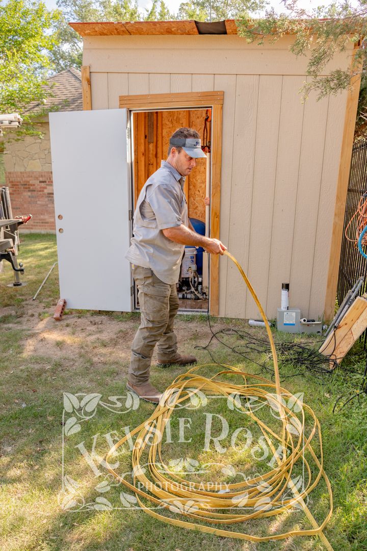 Technician pulling well pump by coiling yellow cable outside well building in backyard.