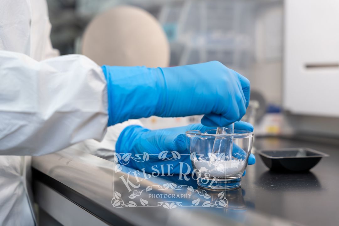 Close-up of gloved hands mixing pharmaceutical compounds in a glass beaker in a pharmacy lab.