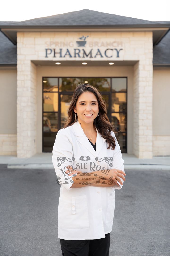 Female pharmacist in white coat posing confidently outside Spring Branch Pharmacy entrance.