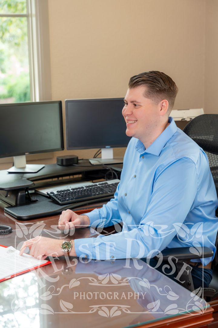 Smiling man in blue dress shirt sitting at office desk with dual monitors and keyboard in professional workspace.