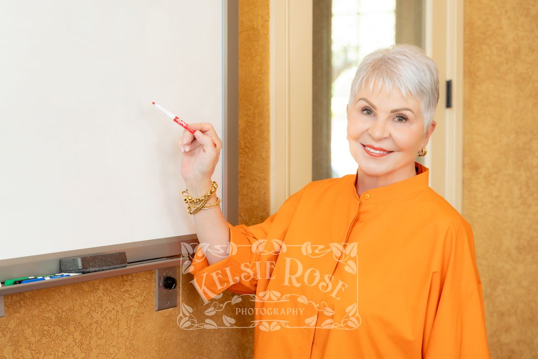 Smiling older woman in orange top writing on a whiteboard, holding a red marker in a bright classroom setting.