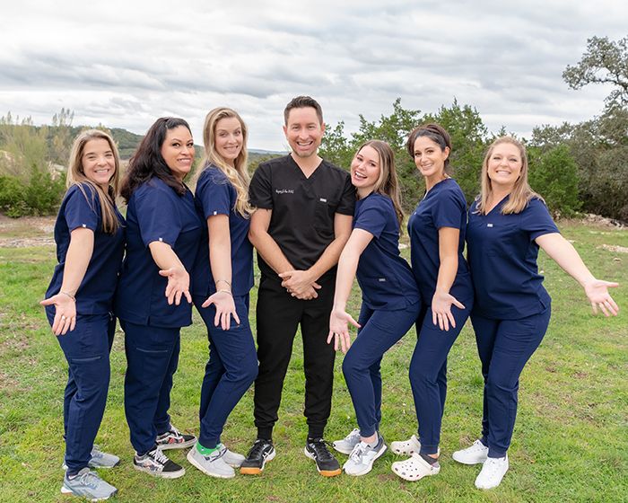 A smiling medical team in navy scrubs poses outdoors in a playful group photo on a green lawn.