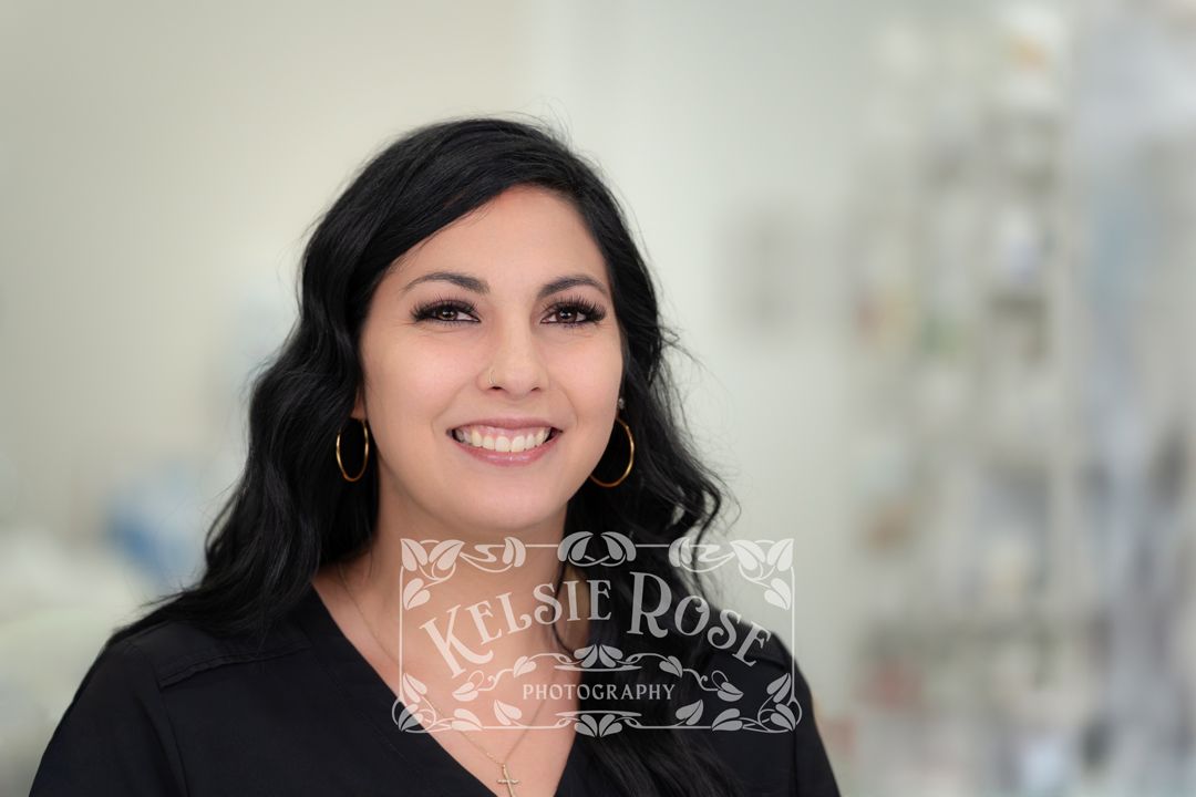 Headshot of smiling dark-haired woman in black top wearing gold hoop earrings in a pharmacy setting.