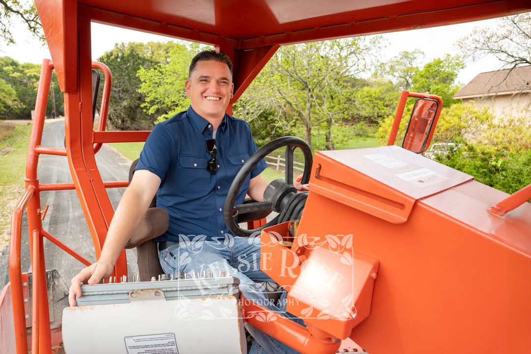 Man in navy shirt smiling while driving and seated in cab of orange road paving equipment.