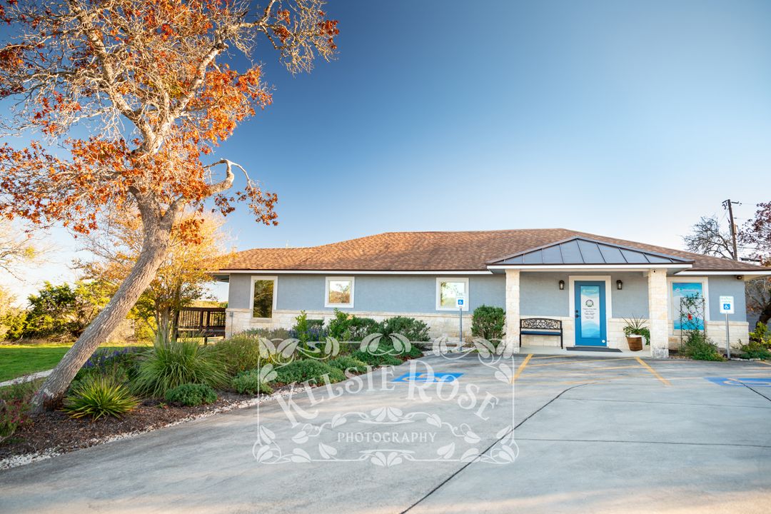 Exterior view of a dental office with a blue door and autumn trees in a sunny parking lot.