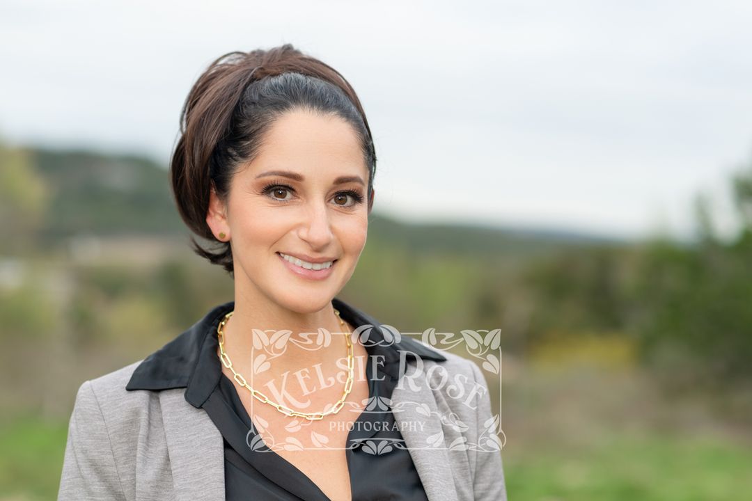 Headshot of professional woman with dark hair in updo wearing gray blazer smiling outdoors with Texas hill country background.