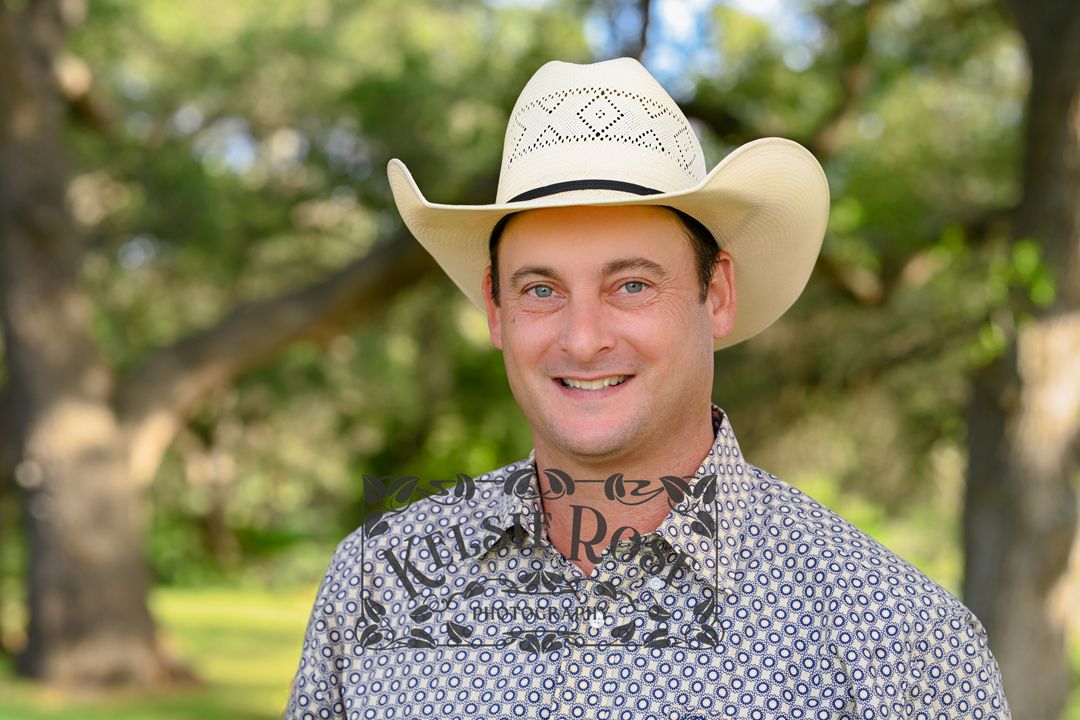 Headshot of smiling man in white cowboy hat and patterned shirt outdoors under trees.