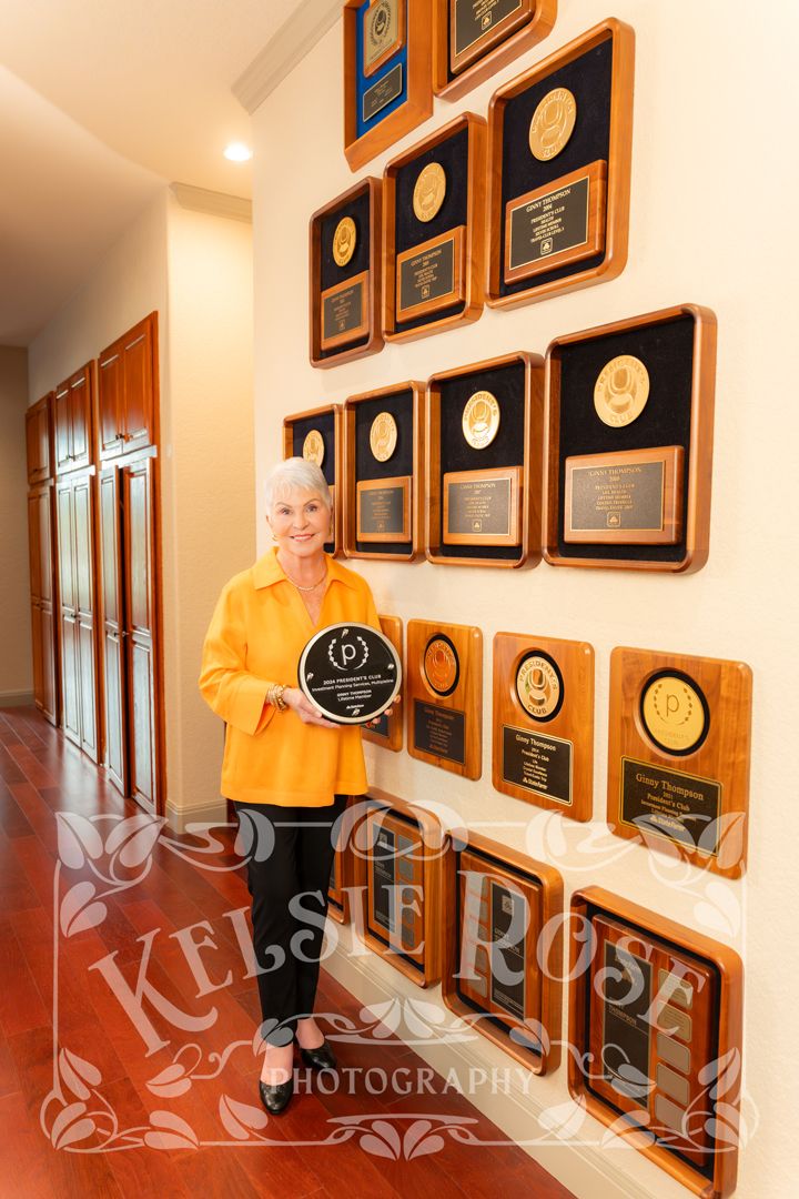 Woman in yellow top proudly holds award plaque beside wall of wooden achievement plaques in hallway.