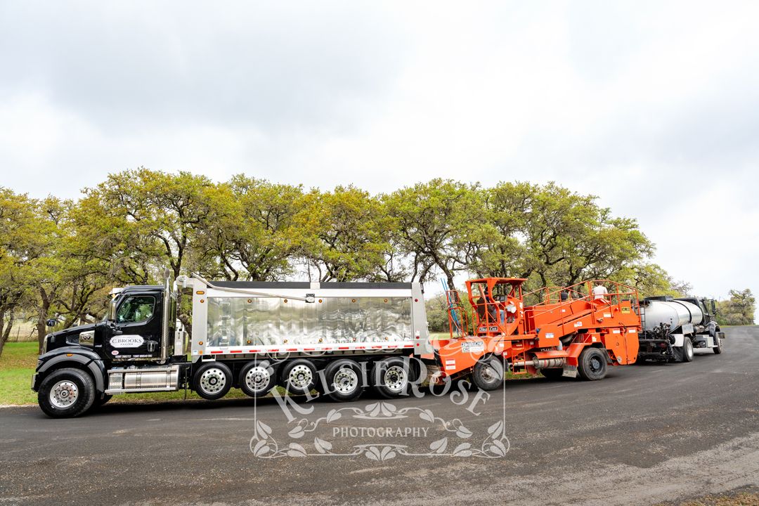 Large dump truck and paving equipment lined up on a freshly paved road surrounded by oak trees.