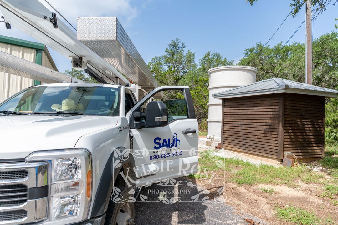 Water well service companty truck with crane parked near a water storage tank and well building.