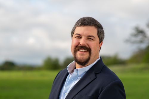 Professional headshot of smiling bearded businessman in navy blazer and light blue shirt poses outdoors.