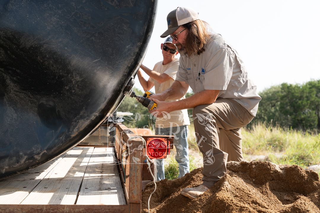Two workers installing a large black pipe in a trench on a sunny construction site outdoors.