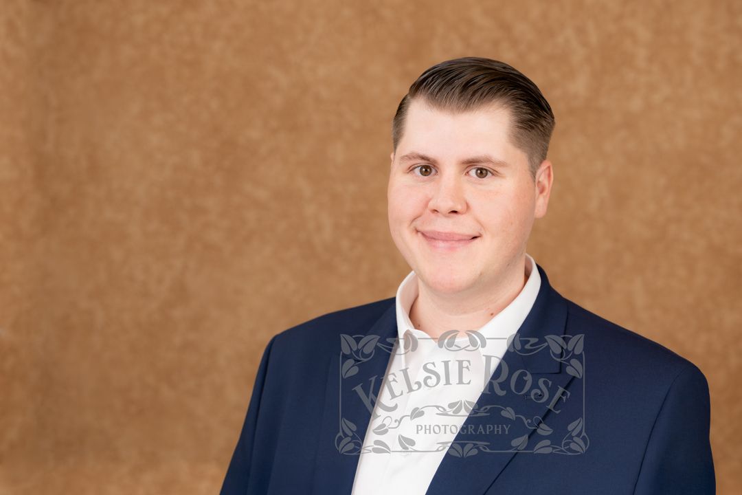 Professional headshot of a smiling man in a navy blue blazer against a brown studio background.