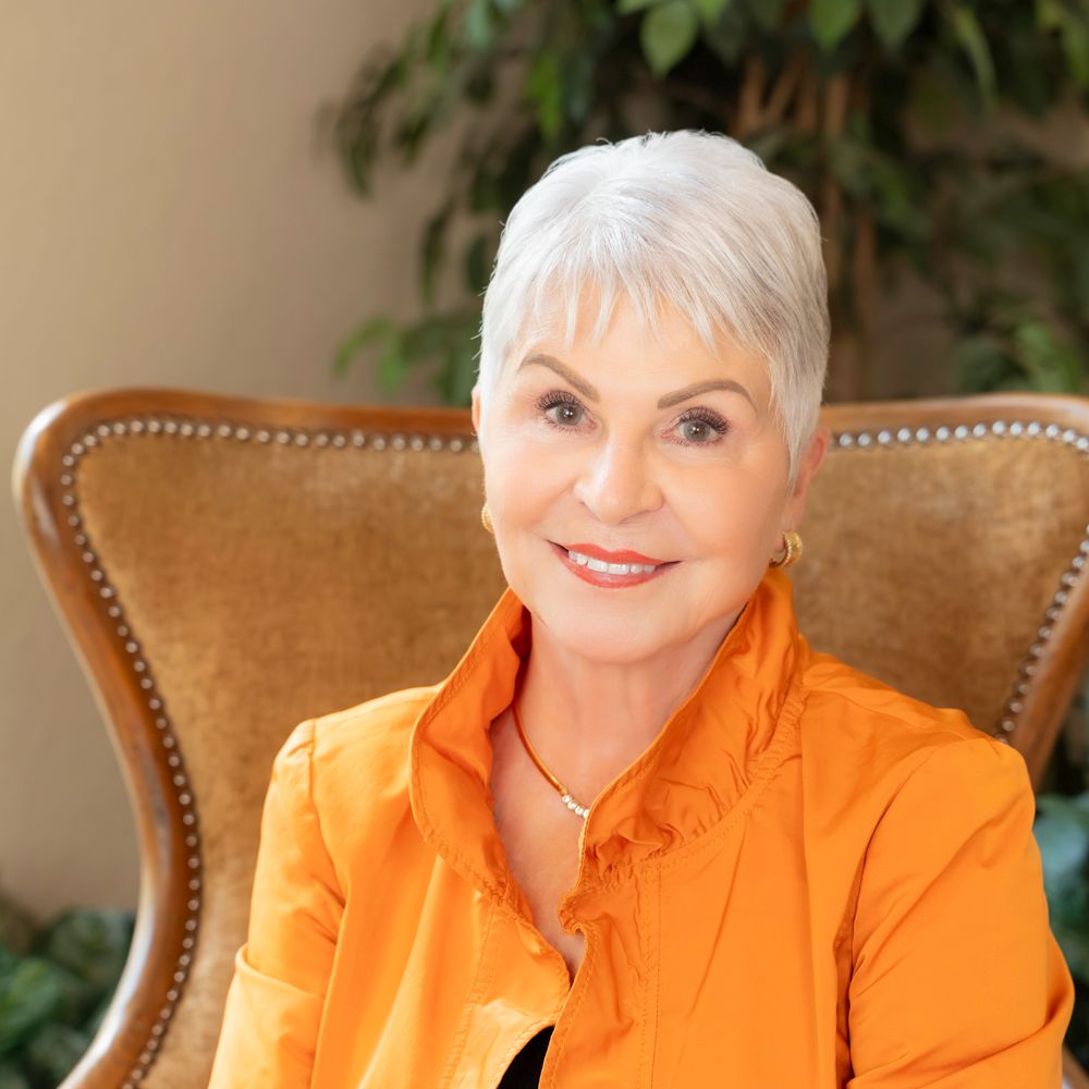 Elegant senior business woman with white hair wearing an orange blouse, smiling warmly while seated in a brown chair.