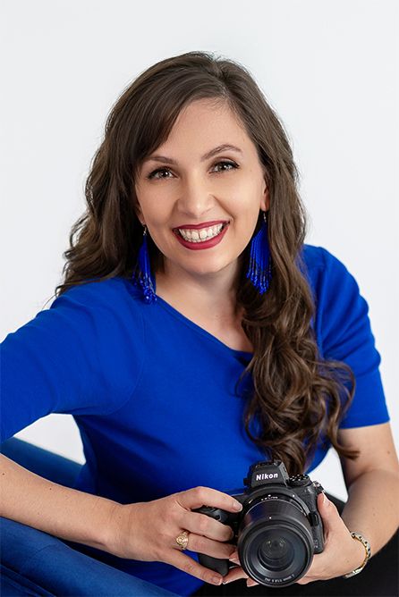 Headshot of smiling female photographer in royal blue top and blue earrings holding camera for professional portrait.