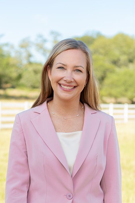 Professional woman in a pink blazer smiling outdoors with a white fence and green field behind her.