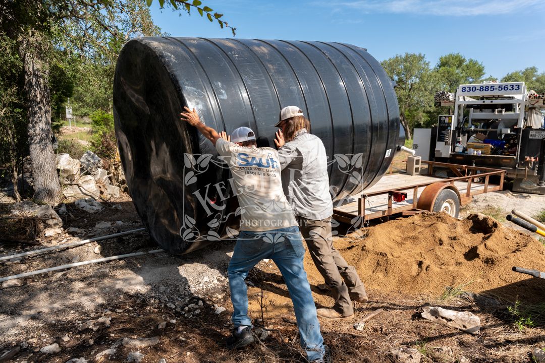 Two workers installing a large black corrugated metal culvert pipe outdoors near trees and a trailer.
