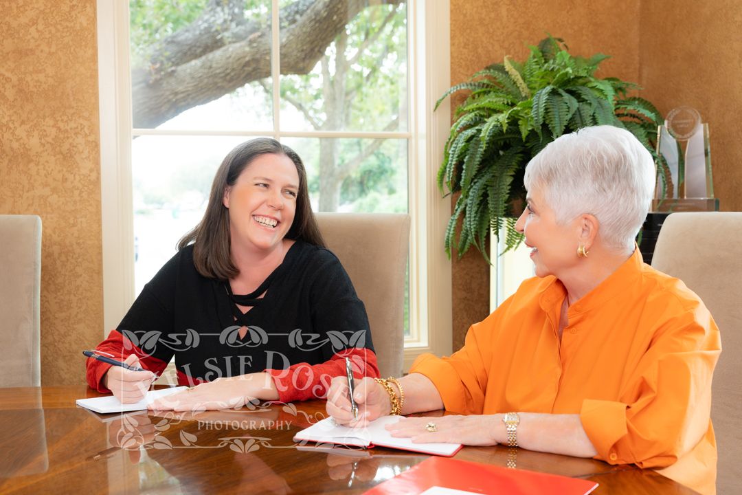 Two women smiling and signing documents together at a wooden table in a bright, elegant room.