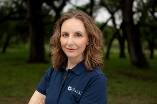 Professional headshot of businesswoman in navy blue polo shirt with arms crossed smiling outdoors among trees.