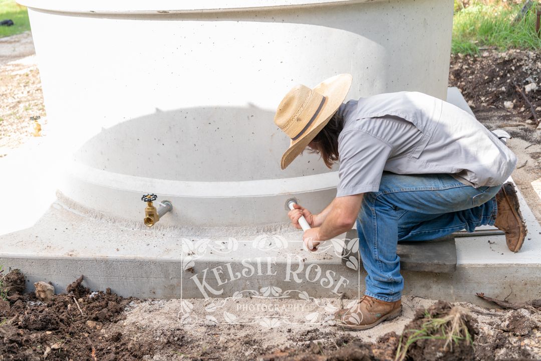 Worker in cowboy hat crouching to install a PVC pipe on a concrete water storage tank base.