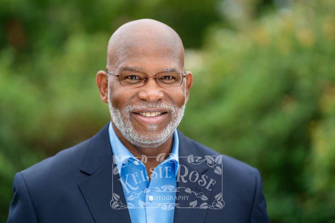 Headshot of smiling middle-aged man with glasses and gray beard wearing a navy blazer outdoors with green foliage background.