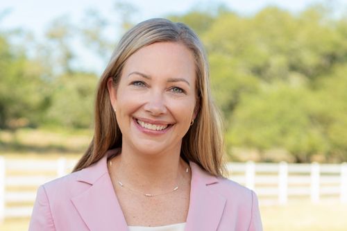 Headshot of smiling blonde woman in pink blazer posing outdoors near white fence and green trees in professional headshot.