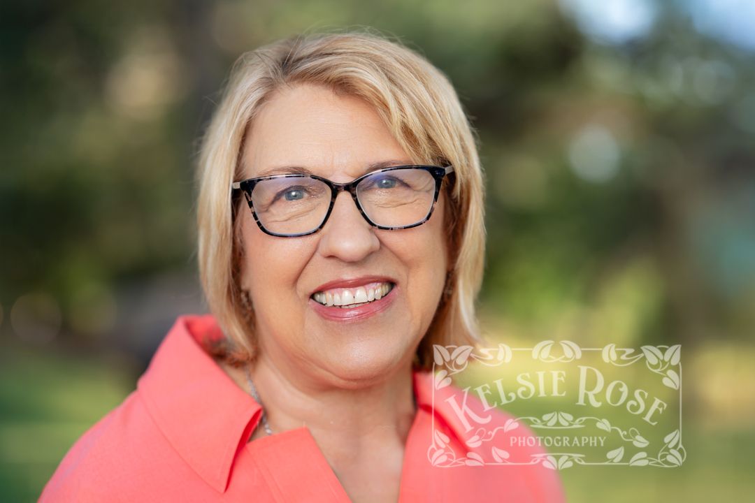 Headshot of smiling middle-aged woman with blonde hair and glasses wearing a coral shirt, outdoor portrait.