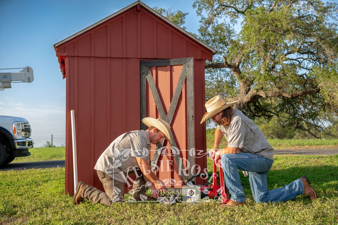 Two workers in cowboy hats kneeling with tools at red well building on green lawn.
