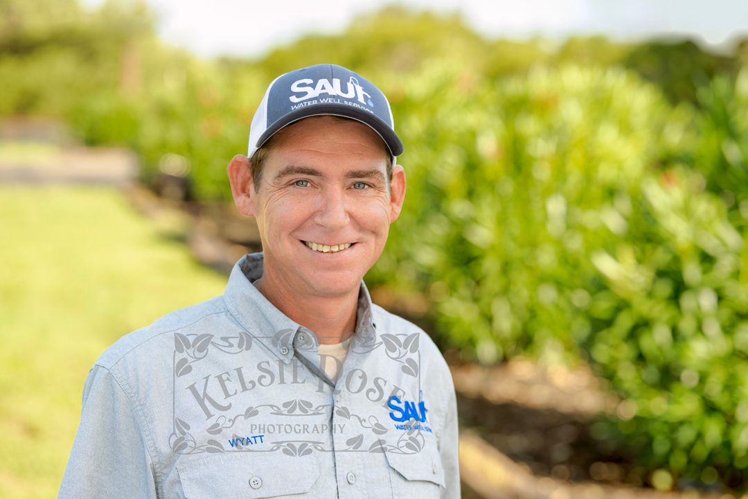Smiling man wearing a branded cap and shirt stands outdoors near oleander bushes on a sunny day in a rural Texas setting.
