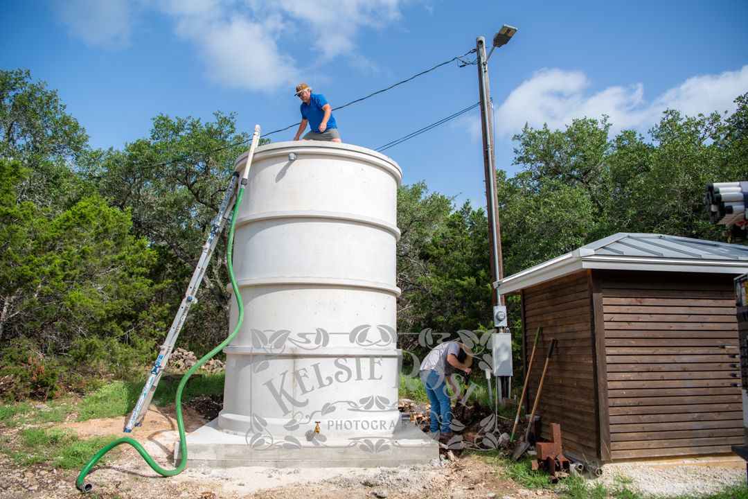 Two workers filling a large white concrete water storage tank with water outdoors near a wooden well building.