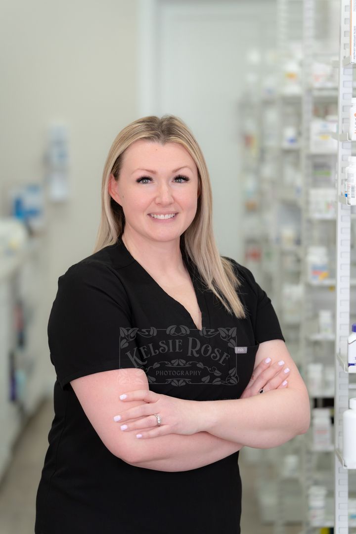 Confident blonde female pharmacy technician in black scrubs posing with arms crossed in a pharmacy.
