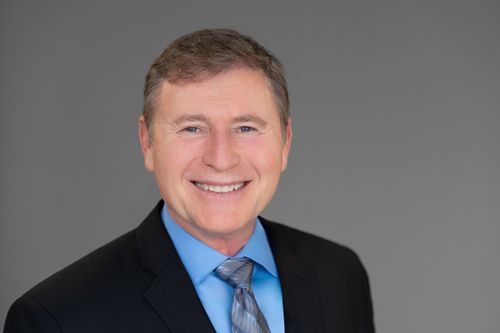 Professional headshot of a smiling middle-aged man in a dark suit and blue tie against a gray background.