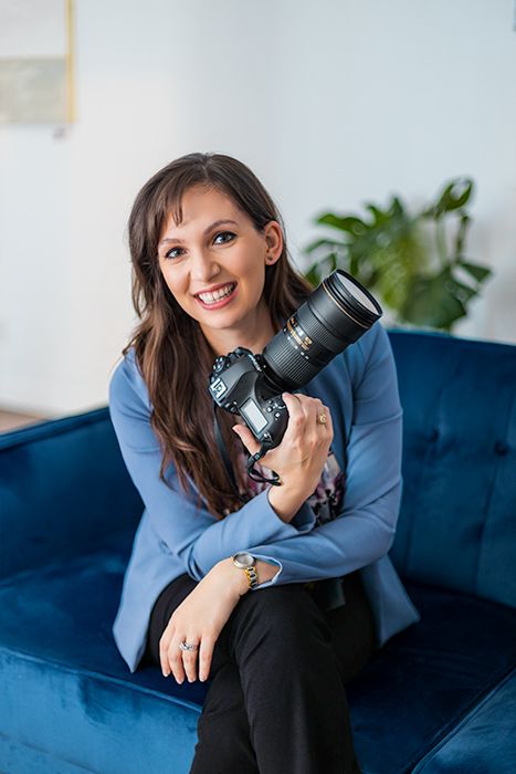 Portrait of smiling female photographer holding DSLR camera with telephoto lens seated on blue velvet sofa in a brightly lit room.