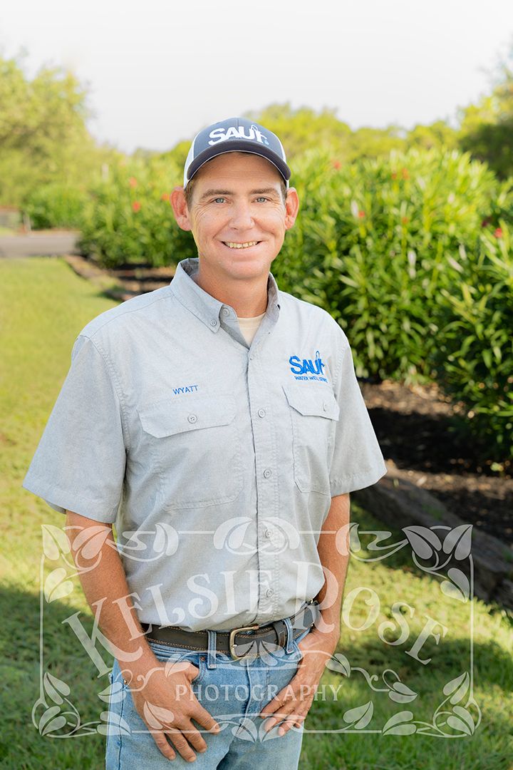 Man in branded shirt and cap smiling outdoors in a green landscaped setting.