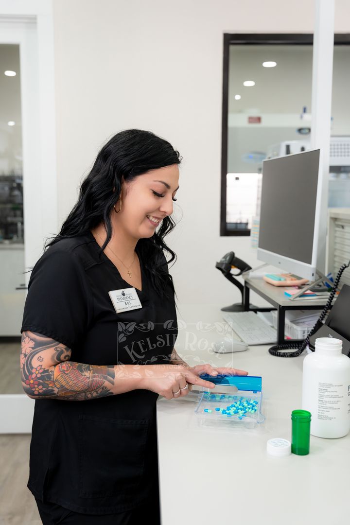 Pharmacy technician in black scrubs sorting blue and aqua capsules at a pharmacy counter.