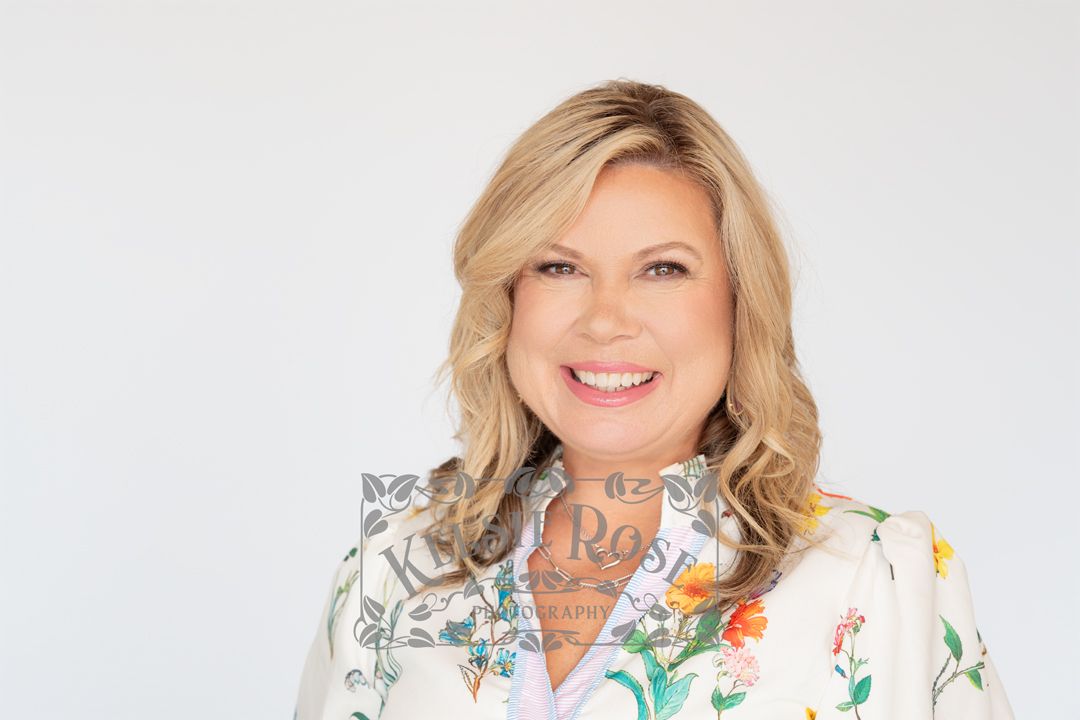 Smiling blonde woman in a floral white dress poses for a professional headshot against a white background.