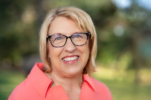 Professional headshot of a smiling businesswoman with blonde hair and glasses wearing a coral shirt outdoors.