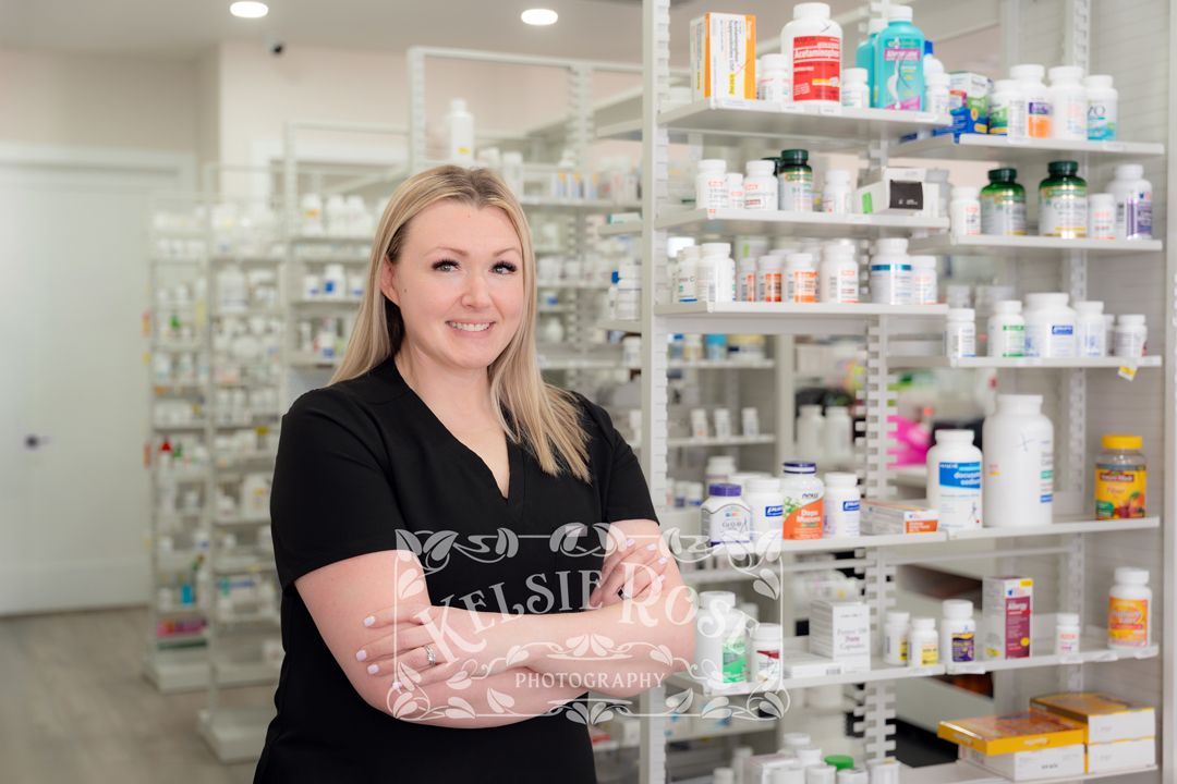 Smiling blonde female pharmacy technician in black scrubs standing with arms crossed in a pharmacy.