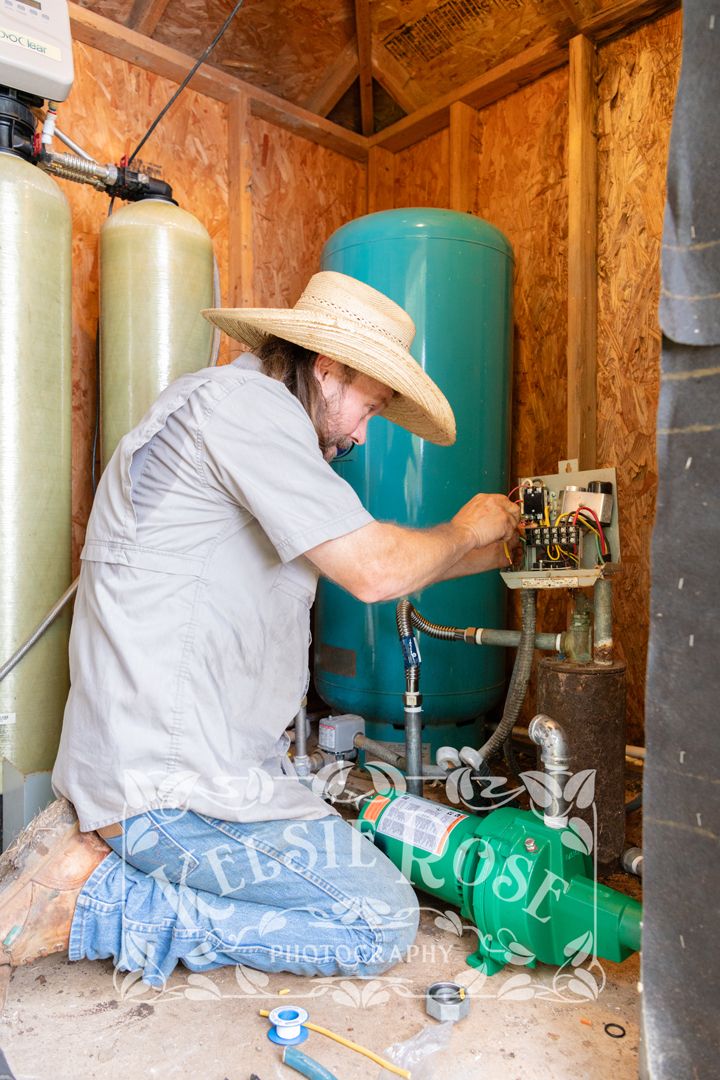 Technician in straw hat working on electrical pump system inside a wooden well building.