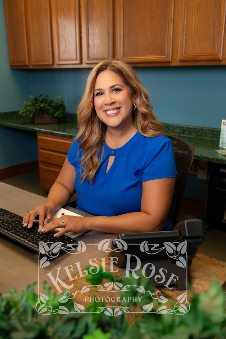 Professional woman in blue blouse smiling at desk with computer keyboard in an office setting.
