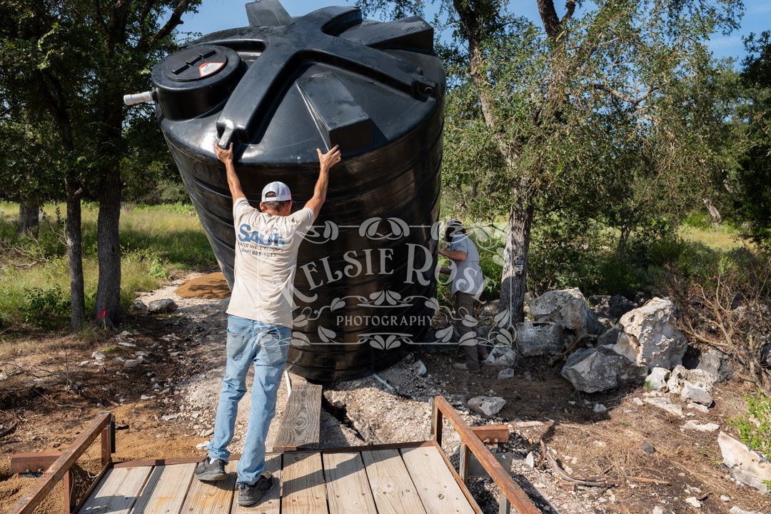 Worker unloading a large black water storage tank from a trailer outdoors on a sunny day.