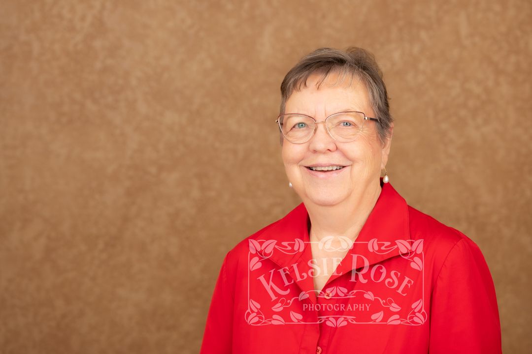 Smiling elderly woman with glasses wearing a red jacket, photographed against a brown studio background.