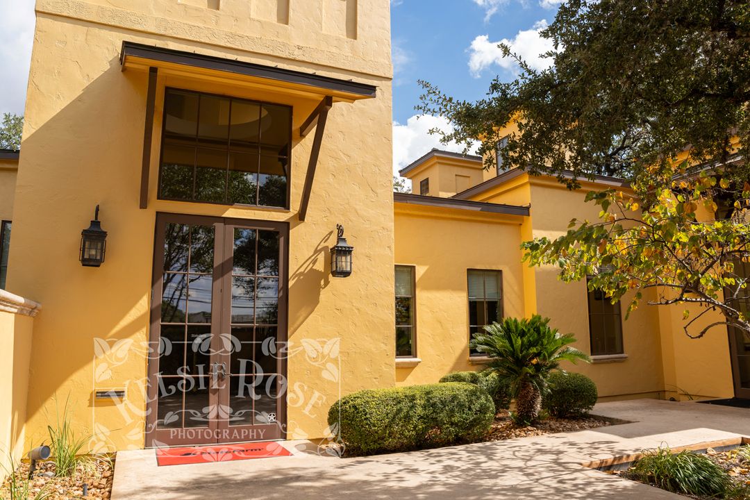 Yellow stucco commercial building entrance with large windows, lantern sconces, and tropical landscaping in Texas.