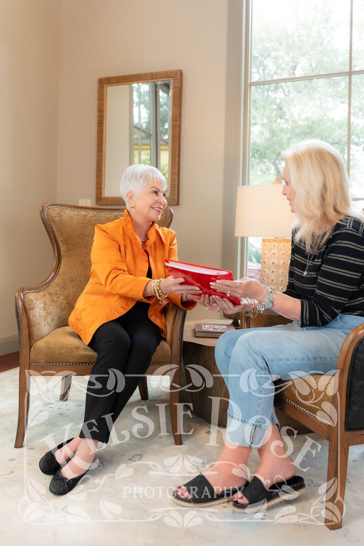 An older woman in an orange jacket hands a red book to a blonde woman while seated in a elegant living room.