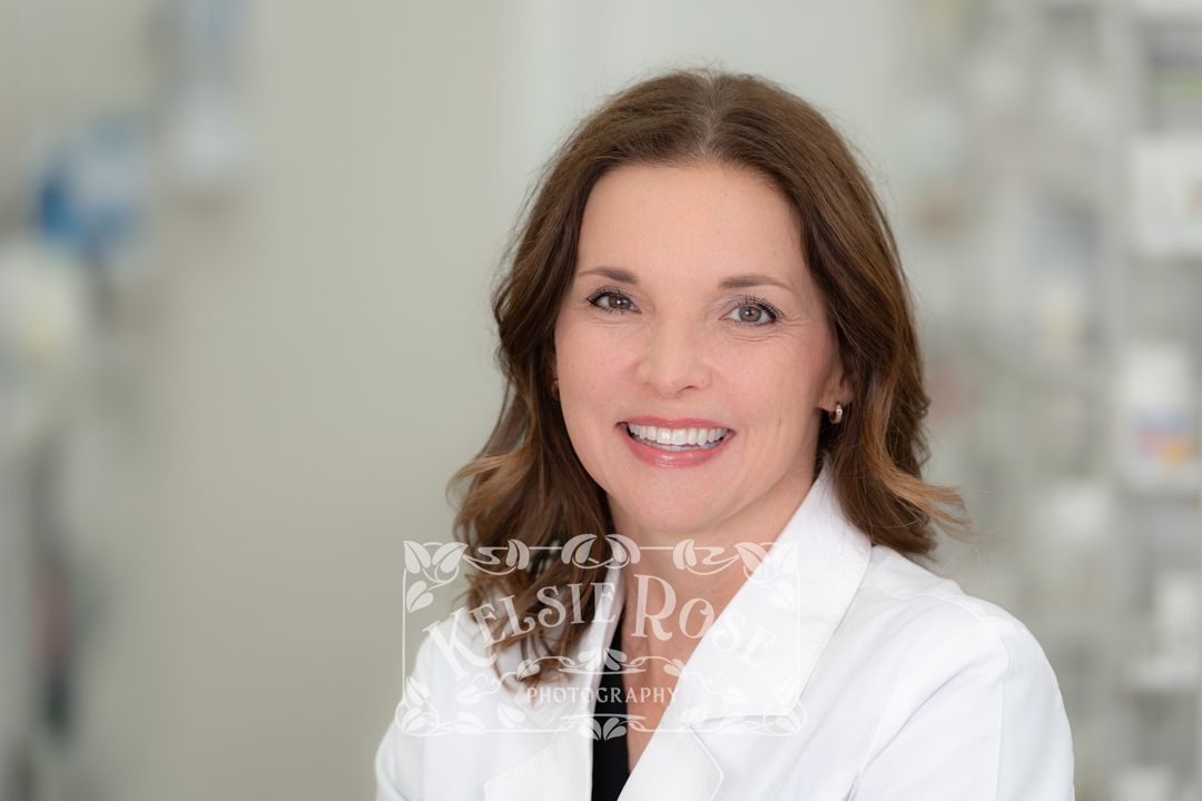 Headshot a smiling female pharmacist in a white lab coat inside a pharmacy setting.