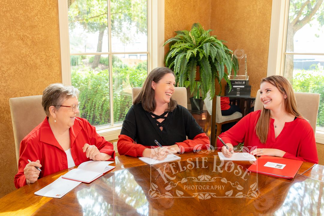 Three women in red and black attire smile while reviewing documents at a wooden conference table near bright windows.
