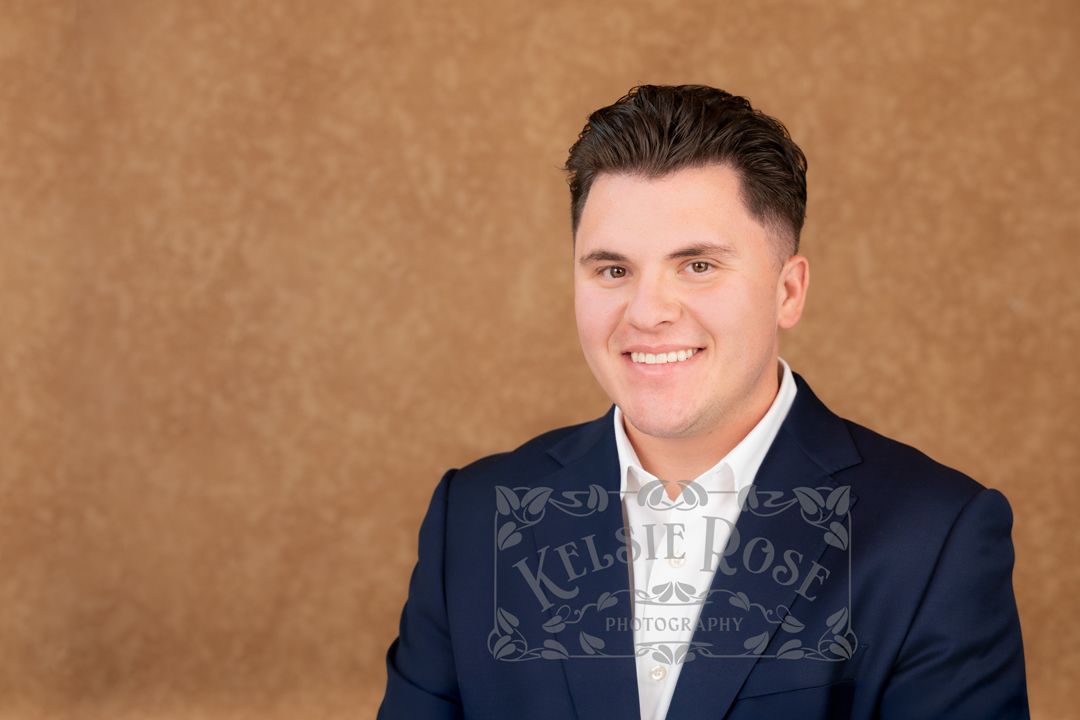 Professional headshot of a smiling man in a navy blazer against a warm brown textured background.